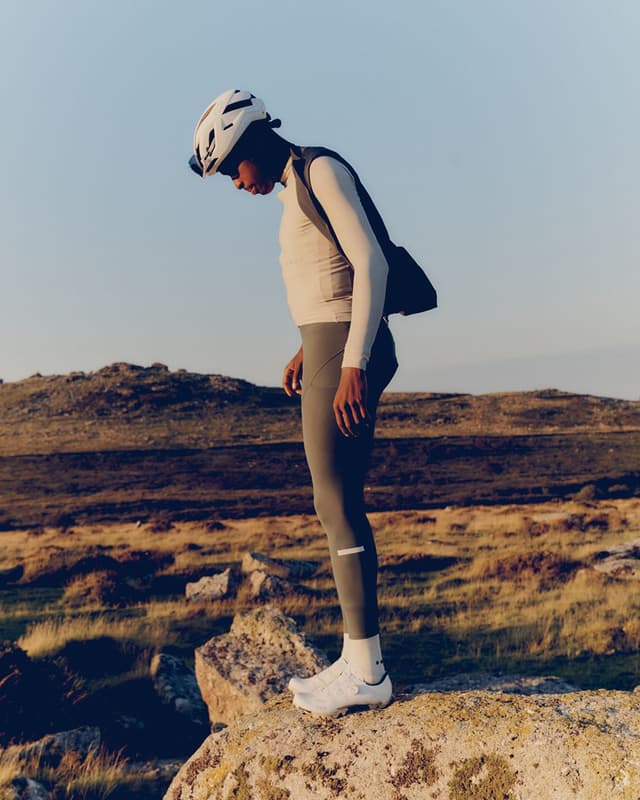A cyclist wearing a white helmet and gear stands on a rock in a rugged landscape, looking down.