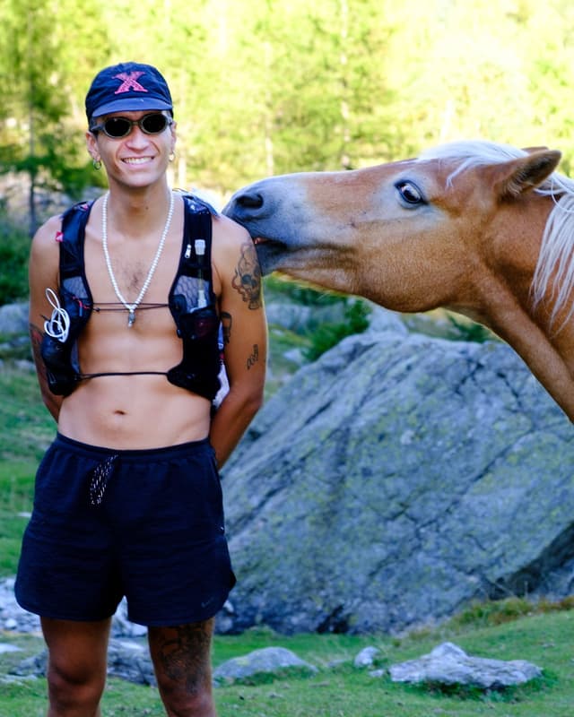 A smiling person in running gear stands outdoors as a horse licks their shoulder.