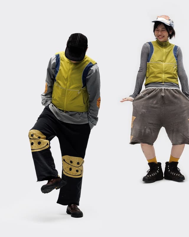 Two people pose in layered outfits: the left wears a black cap, yellow vest, and black pants with yellow smiley knee patches; the right wears a patterned cap, yellow vest, and baggy brown shorts.