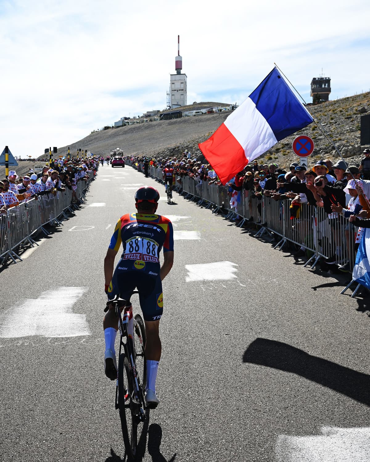 A cyclist climbs a mountain road lined with cheering spectators and a large French flag, towards a summit building with an antenna.