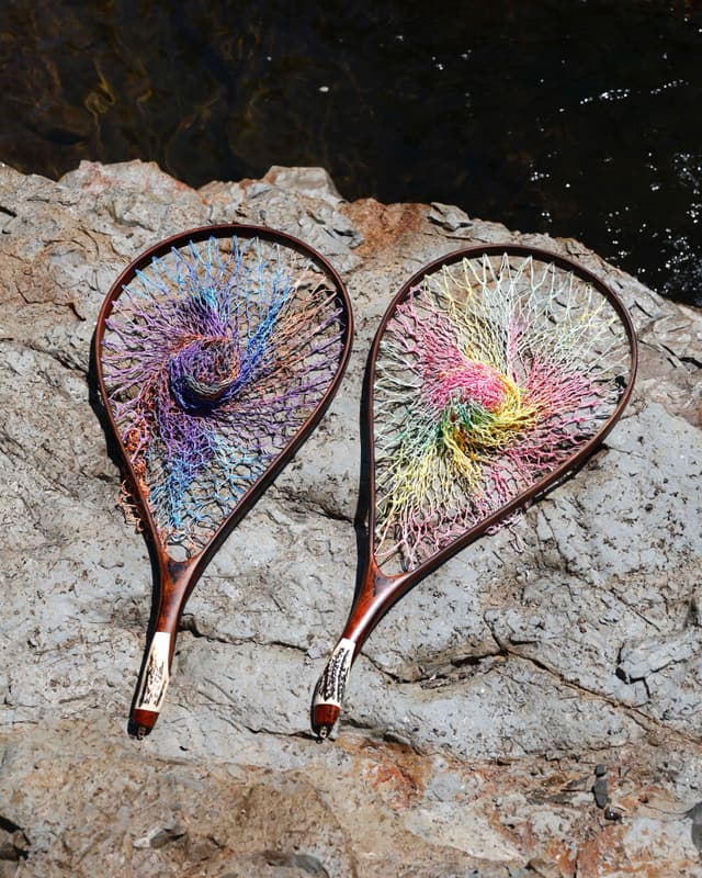 Two ornate landing nets with colorful spiral netting and antler handles on rocks by water.