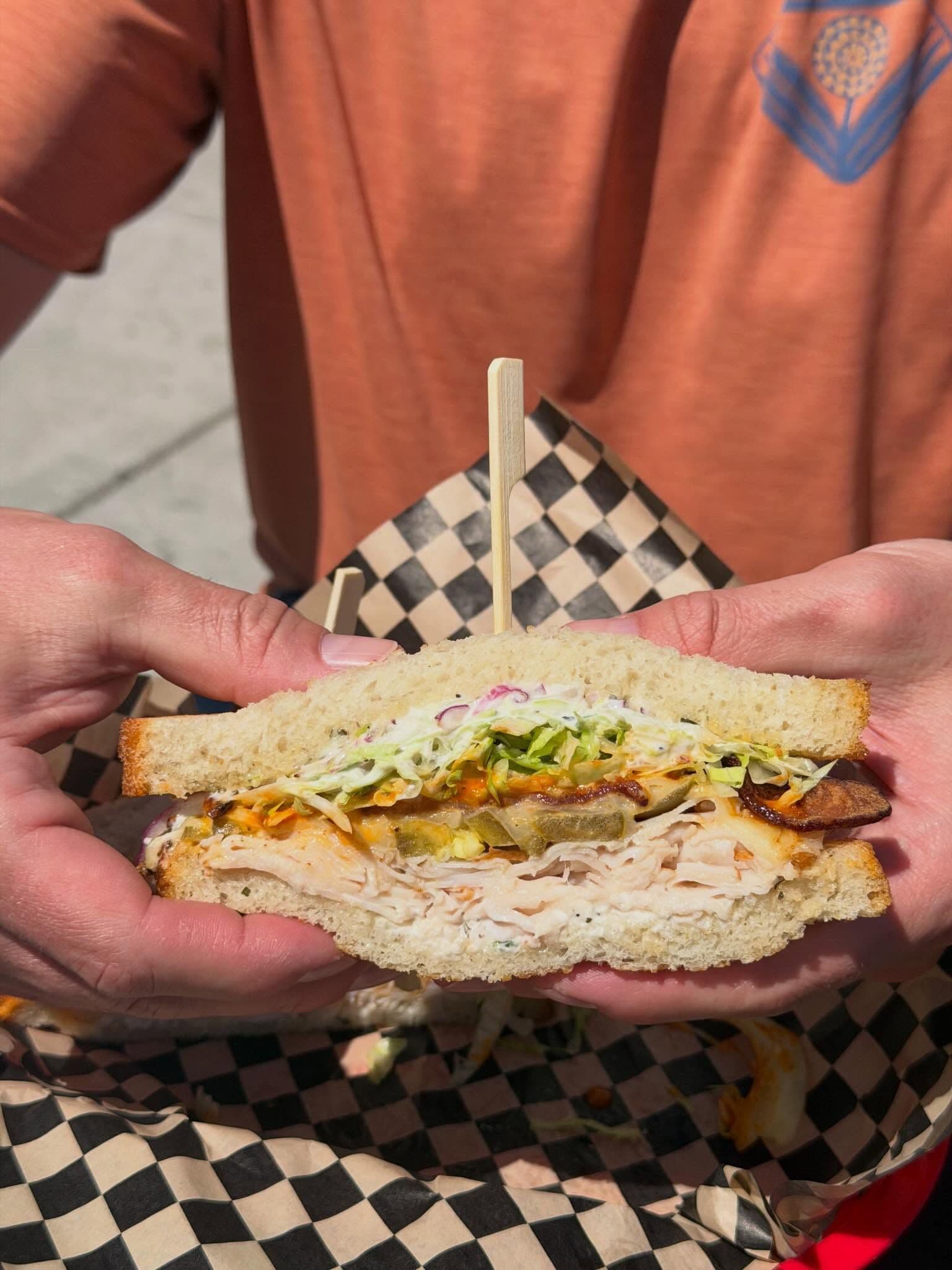 Close-up of hands holding a turkey sandwich with coleslaw and pickles, served in a checkered paper wrapper.