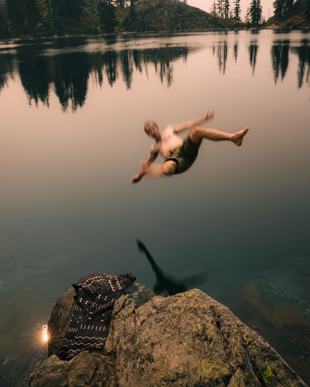 Motion-blurred man jumping into a tranquil lake from a rock, a patterned shirt rests on the rock.