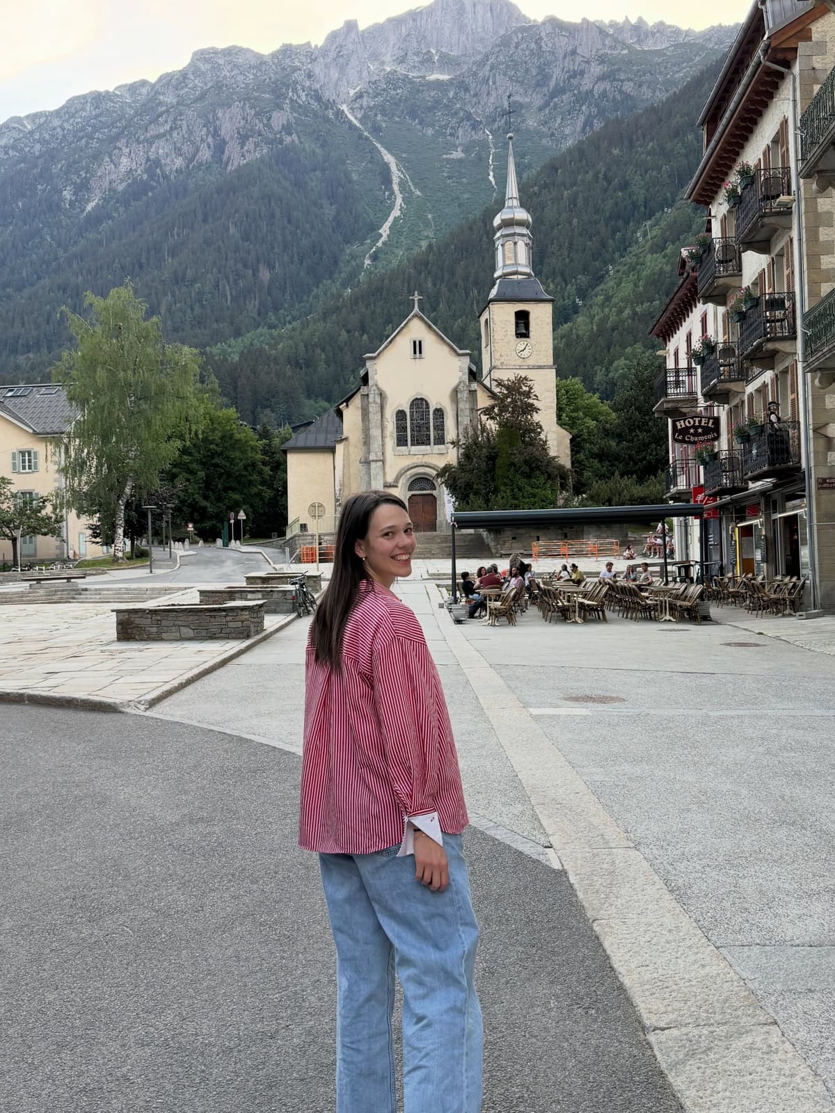 Young woman in a striped shirt smiles over her shoulder on a street with a church and a large, forested mountain in the background.