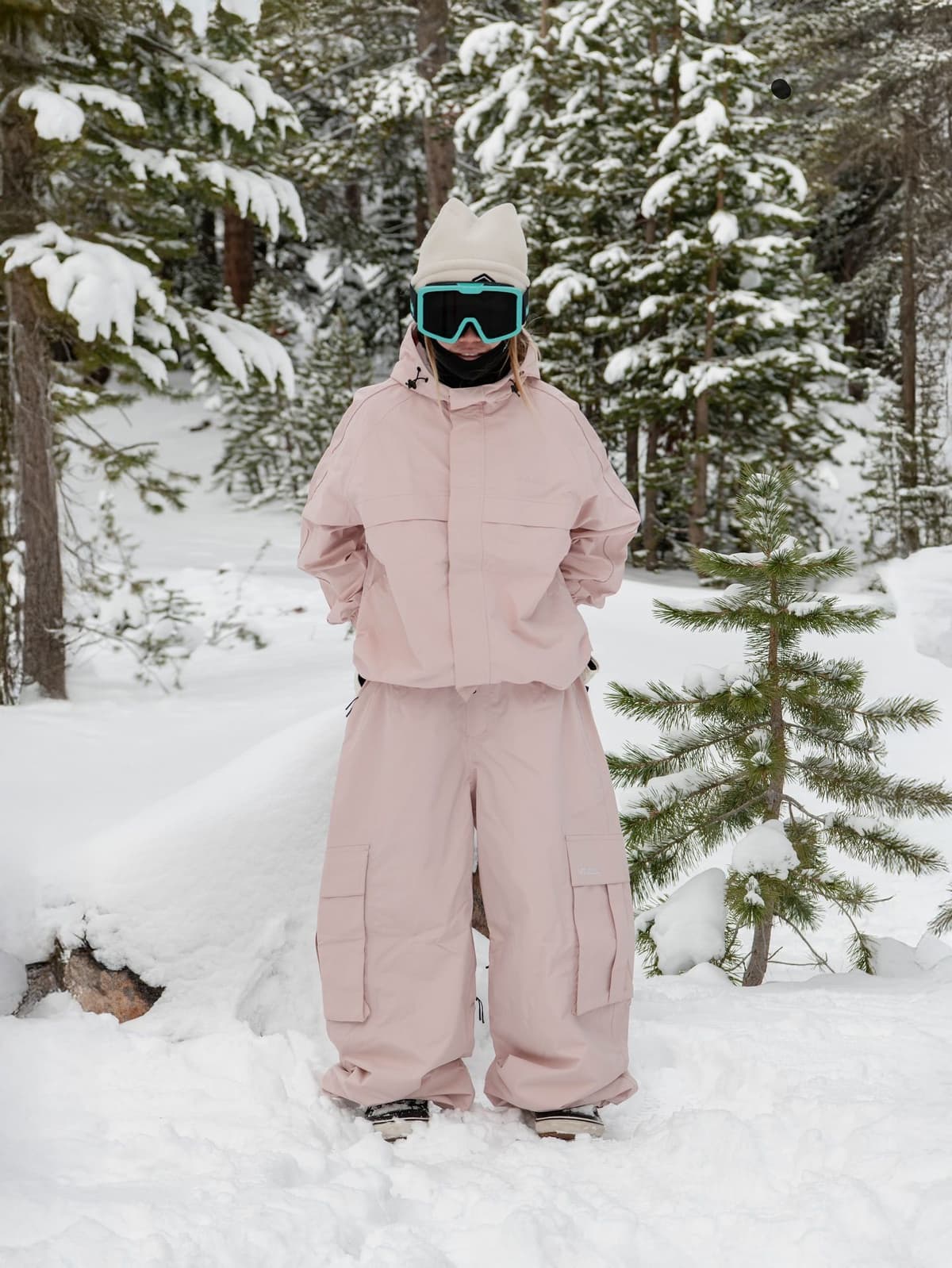 A person in a pink snowsuit, white beanie, and teal goggles stands in a snowy forest.