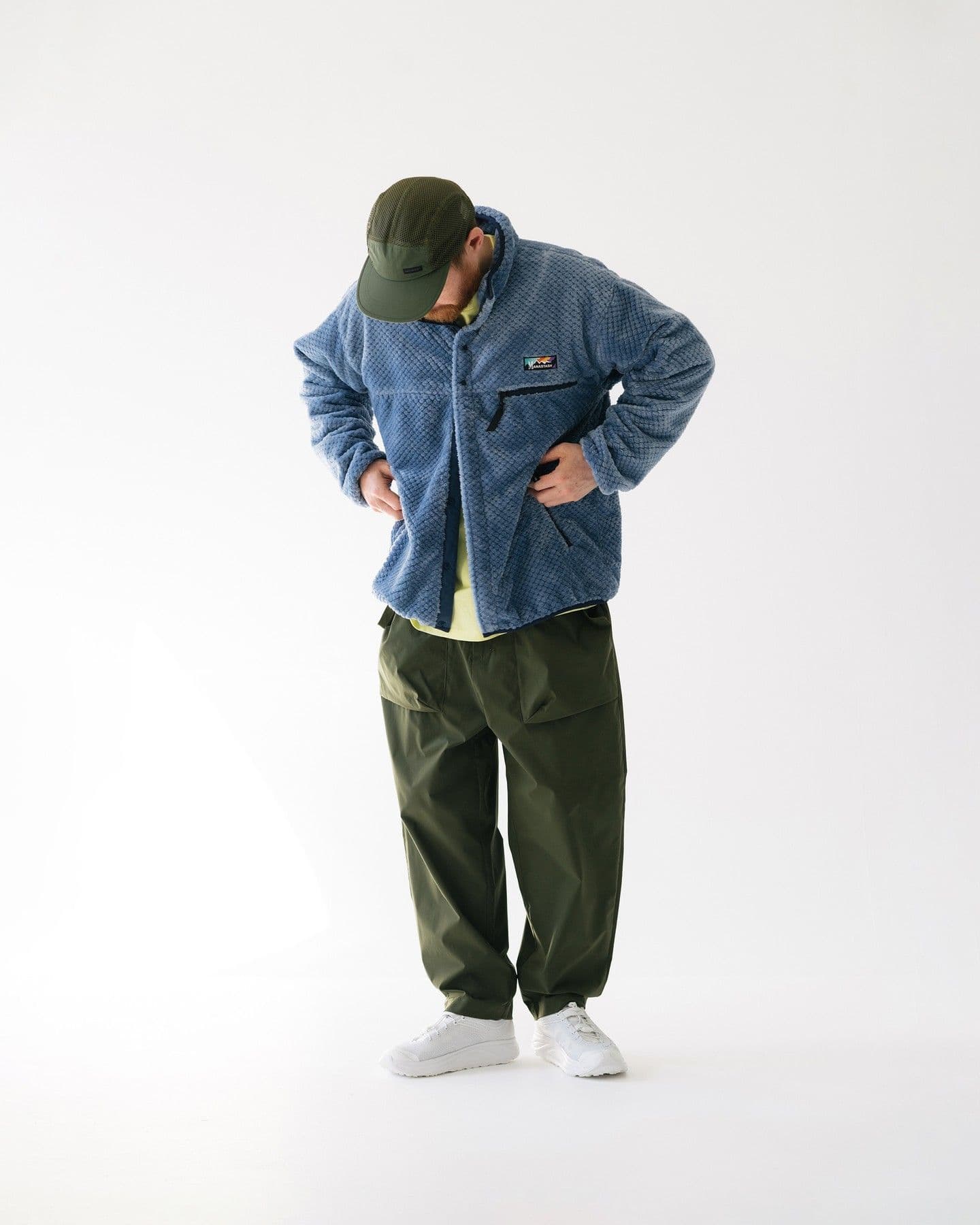 A person wearing a blue fleece jacket, olive green cap and pants, and white sneakers, looking down.