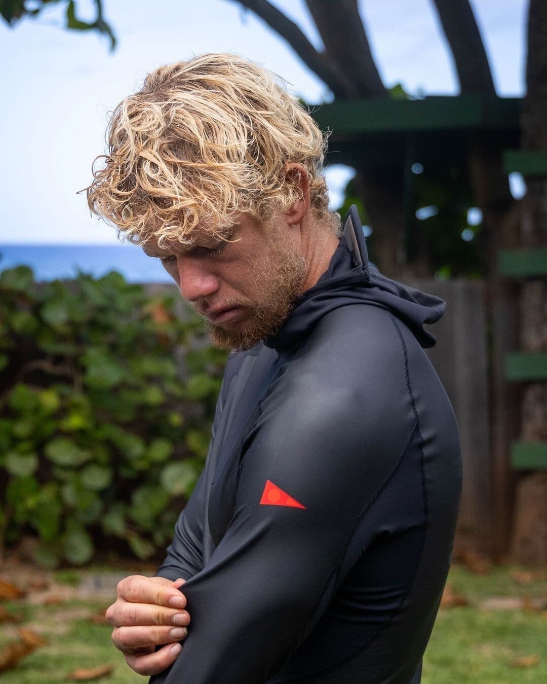 A blonde-haired man with a beard looks down while wearing a dark long-sleeved top with a red triangle logo on the arm, outdoors.