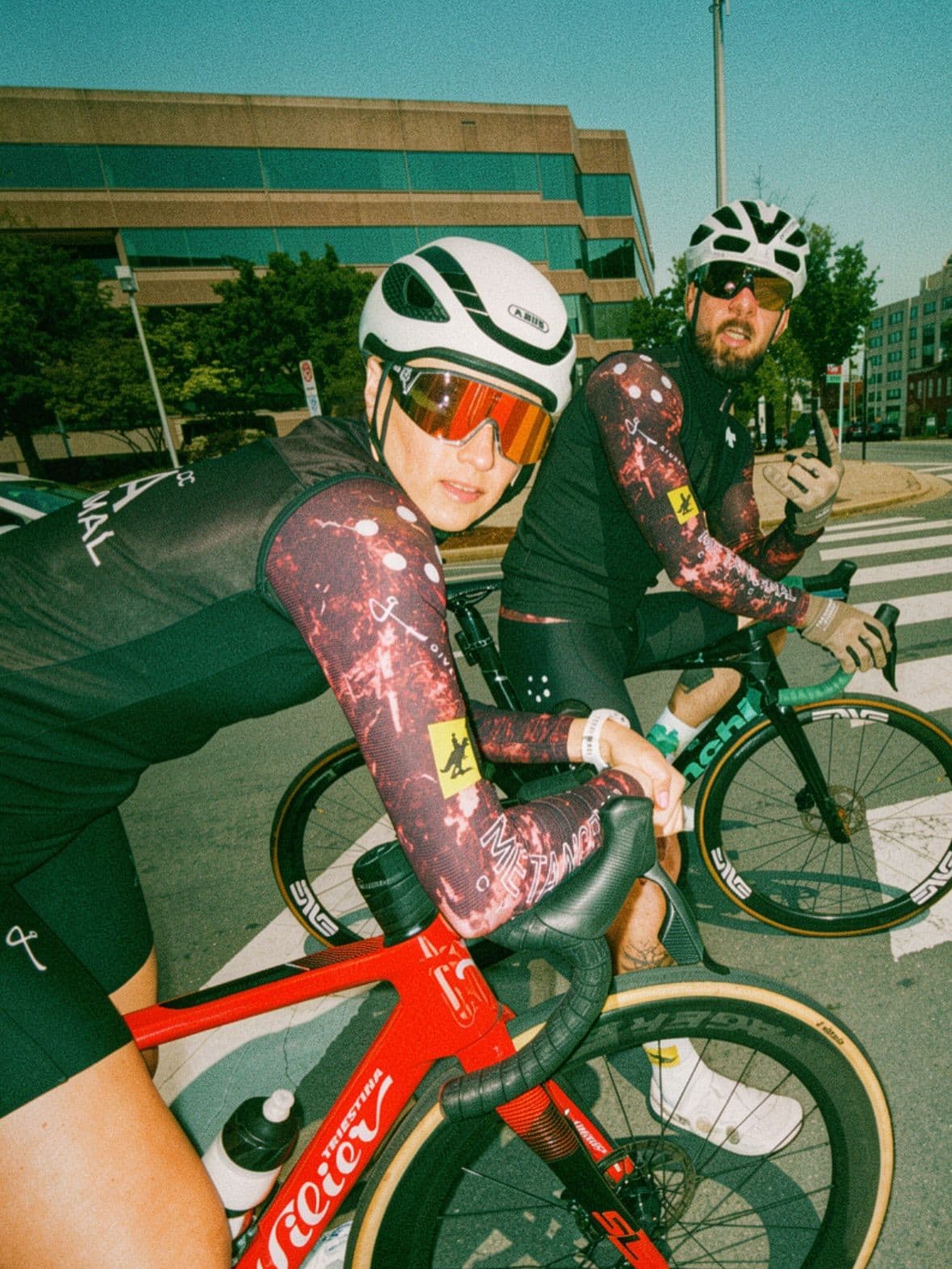 Two cyclists, a woman and a man, wearing helmets and sunglasses, sit on their road bikes on a city street.