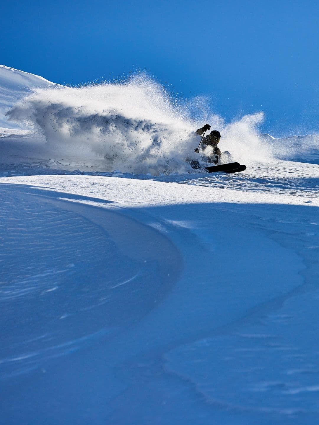 A skier turns sharply, kicking up a large cloud of snow on a sunny, blue-sky day.