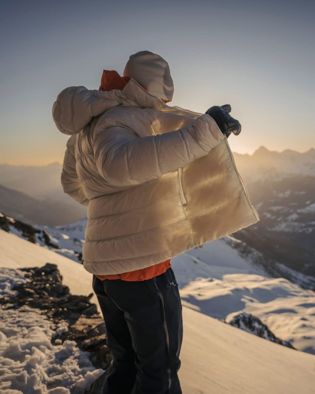 Person zipping up a white puffer jacket on a snowy mountain at sunset.