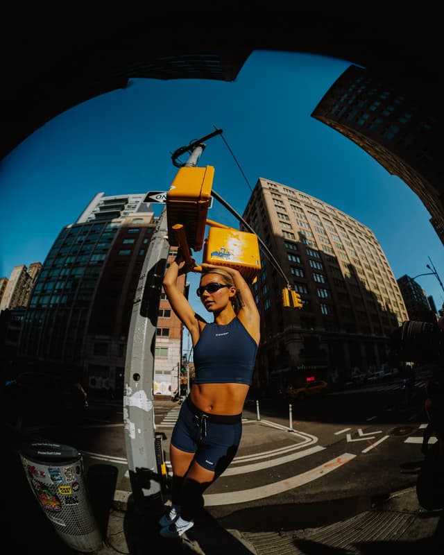 A woman in a navy athletic outfit poses with a traffic light on a sunny city street, fisheye lens perspective.