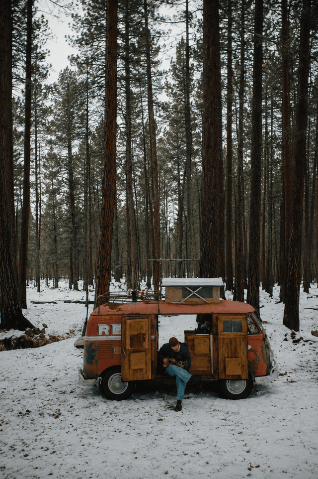 A person plays a guitar while sitting in the open door of a rusty red camper van in a snowy pine forest.