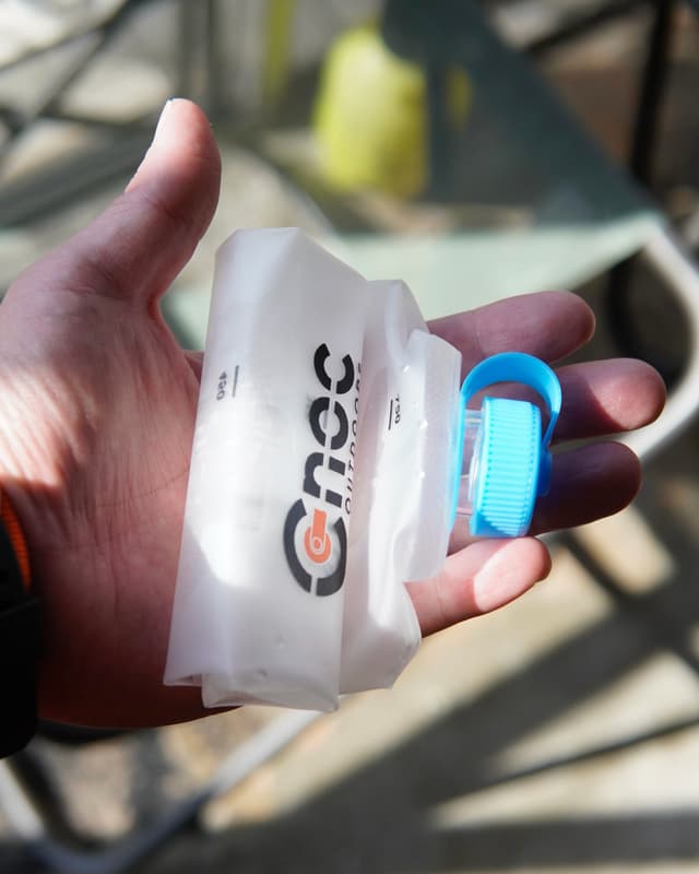 A hand holds a white, collapsible water bottle with a blue cap and "onec outdoors" logo.