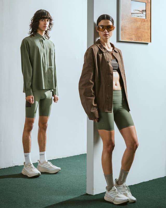 A man in an olive green shirt and bike shorts stands next to a woman in sunglasses, a brown jacket, and olive green bike shorts; both wear sneakers indoors.