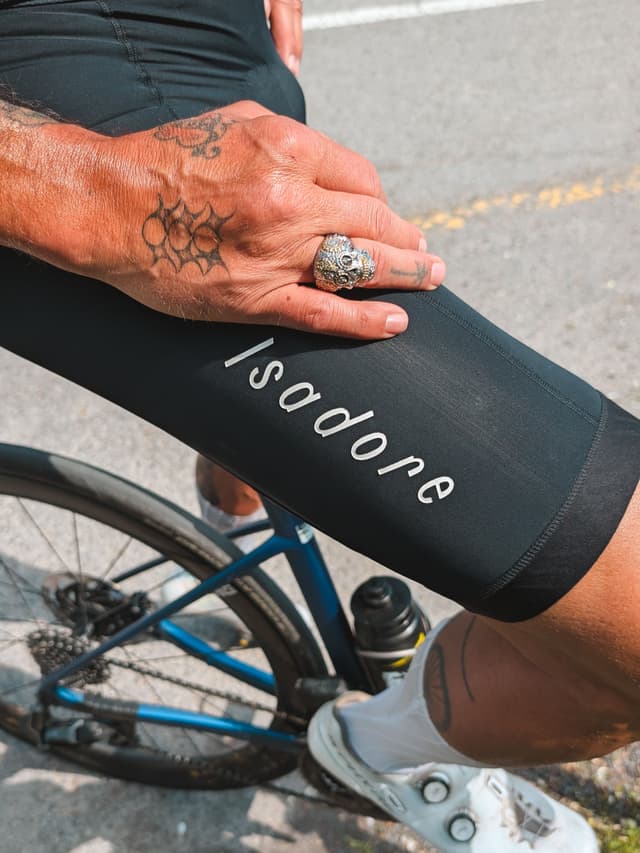 A cyclist's leg in black Isadore shorts, with a tattooed hand wearing a skull ring resting on it, beside a bike.