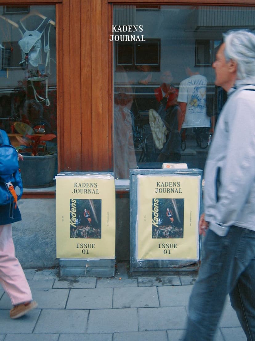 Two "Kadens Journal Issue 01" magazines on sidewalk stands in front of a shop window, with pedestrians walking past.