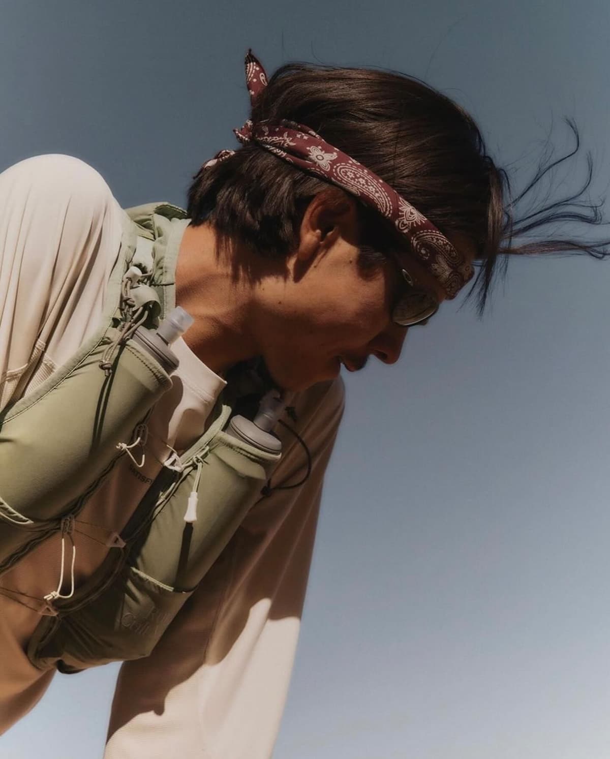 A person wearing a paisley bandana, sunglasses, and a green hydration vest with two water bottles, looks down against a blue sky.