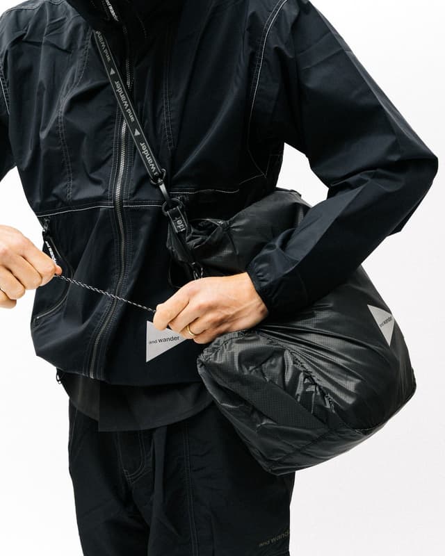A person in a black jacket and pants cinches a black packable bag with "and wander" branding.