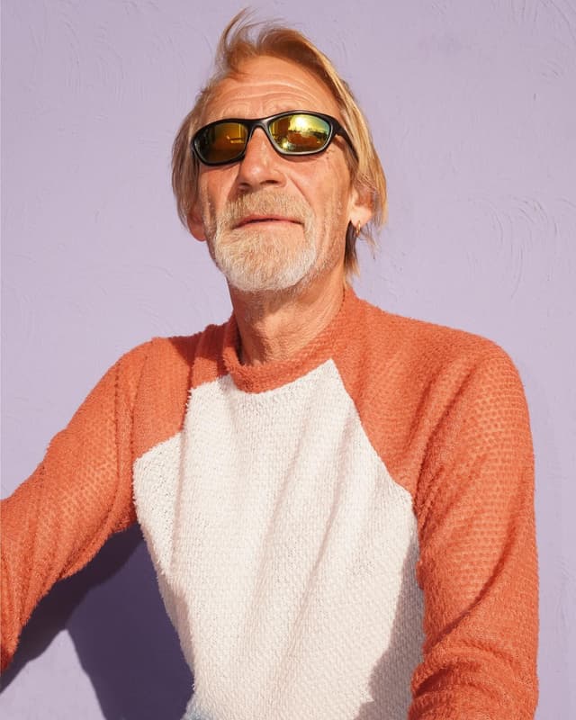 A man with blonde hair and a beard, wearing mirrored sunglasses and an orange and white sweater, against a lavender background.