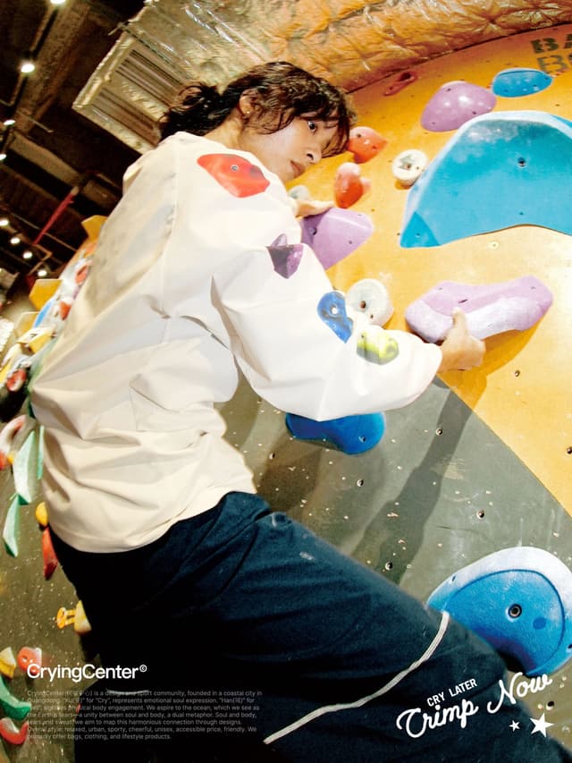 A person with dark curly hair climbs a yellow bouldering wall, reaching for holds.
