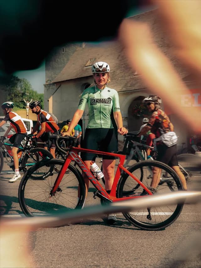A female cyclist in a light green jersey and helmet stands beside a red road bike, with other cyclists and buildings blurred in the background.