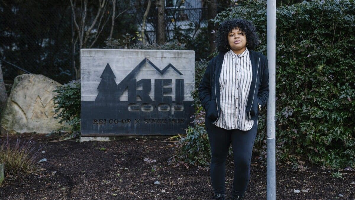 A Black woman with curly hair stands in front of a large REI Co-op sign.