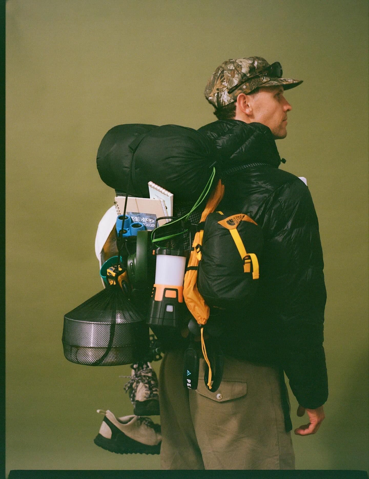 A man in a camo hat and black puffer jacket, viewed from the side, carries a large backpack heavily loaded with camping gear, including a sleeping bag, lantern, and hanging shoes.