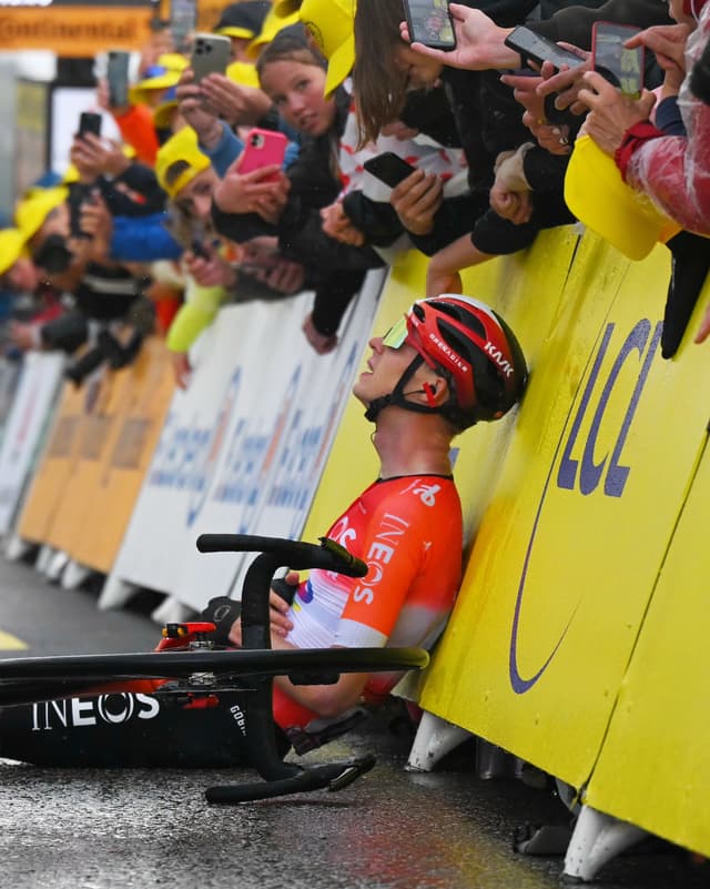 An exhausted cyclist in an orange jersey sits on wet ground by his fallen bike, leaning against a yellow barrier, as a crowd photographs him.