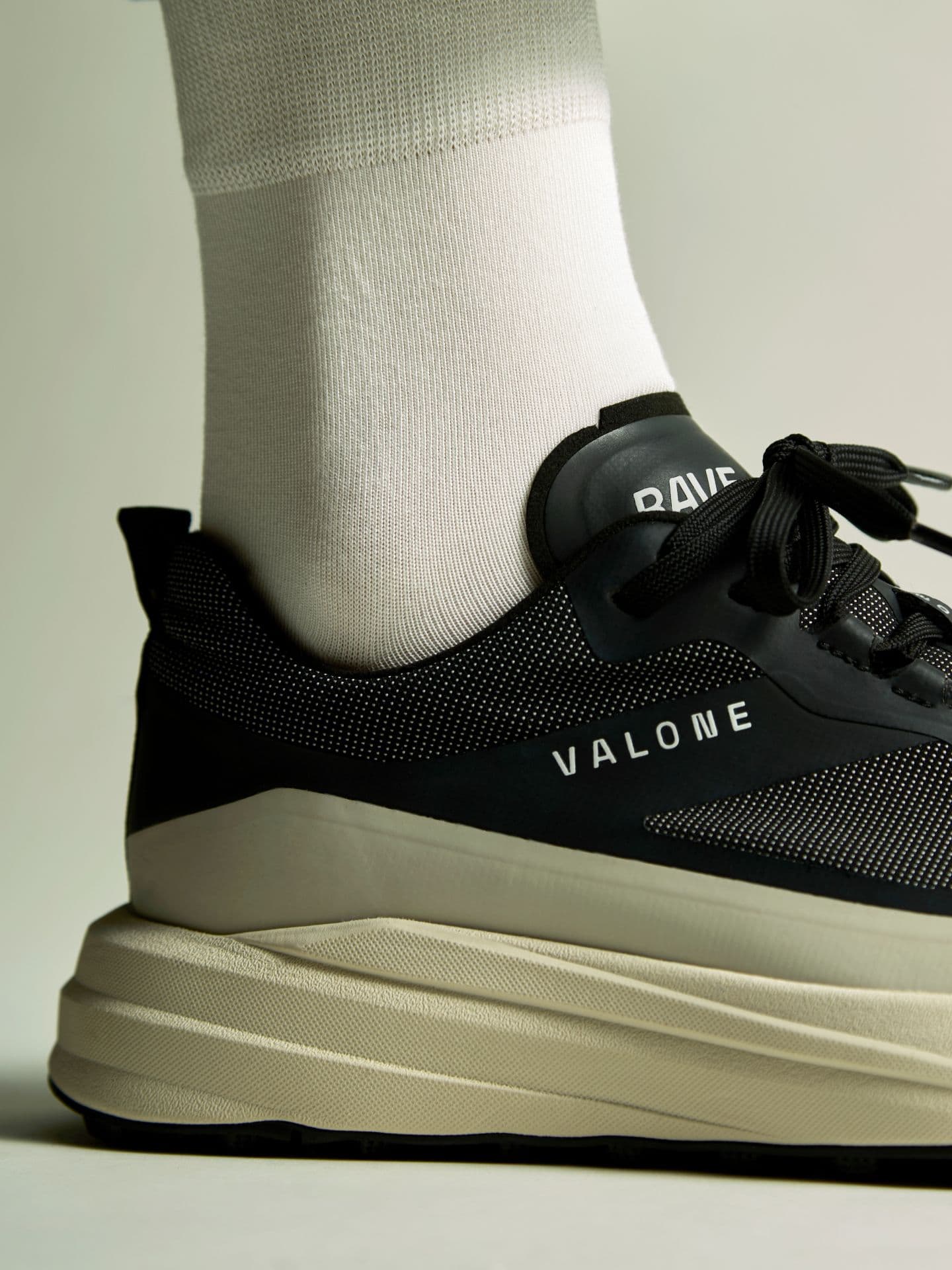 Close-up of a foot in a white sock wearing a black and beige sneaker with a chunky, ridged sole, featuring "VALONE" text on the side.