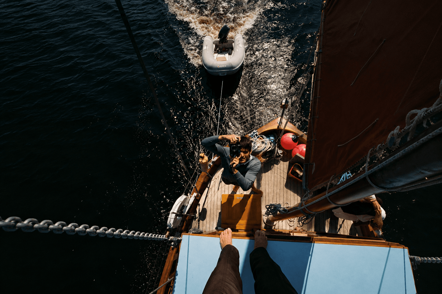 Overhead view of a sailboat deck with a person holding a camera, an inflatable dinghy trailing, and bare feet in the foreground.