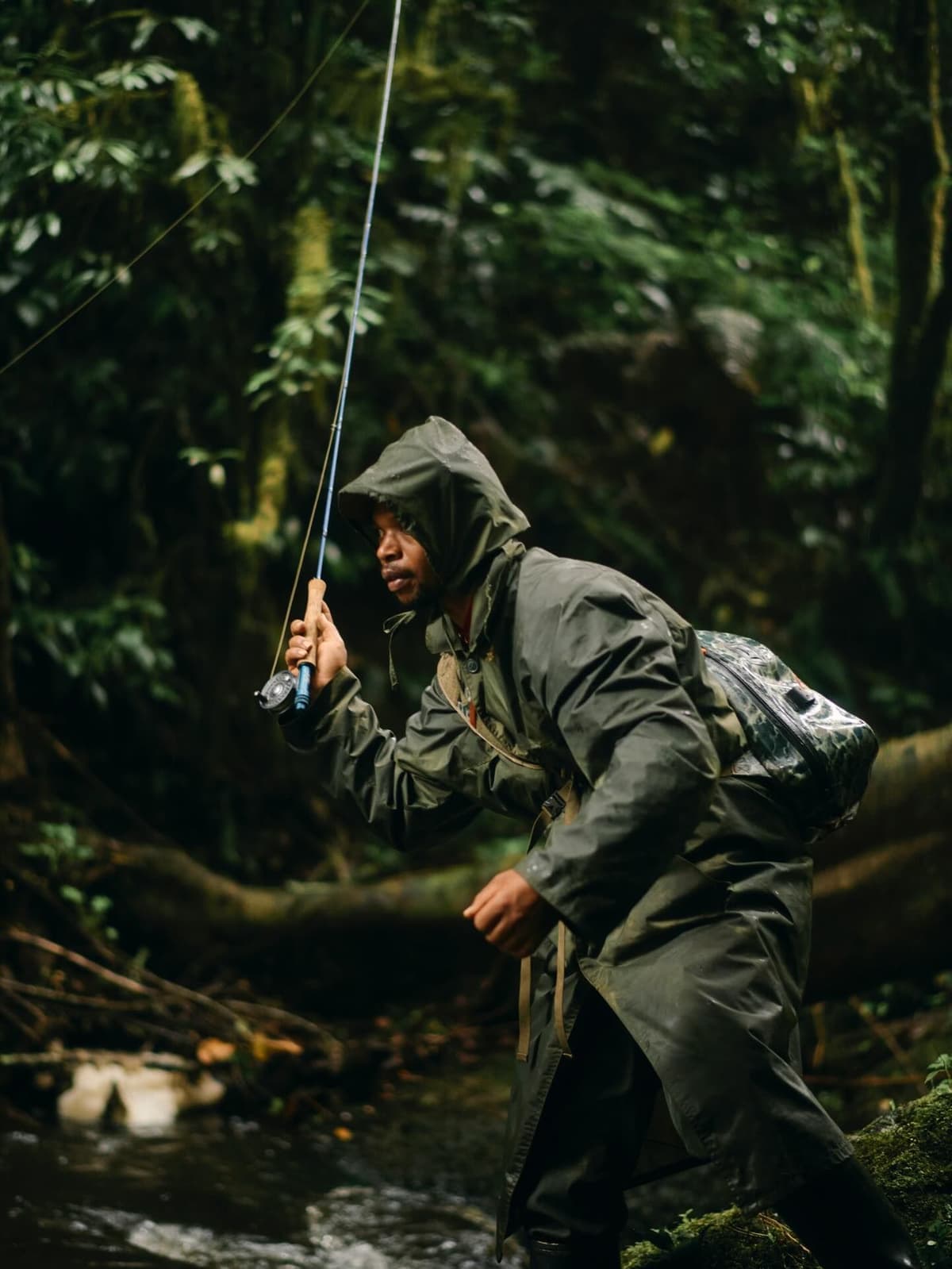 A man in a hooded raincoat fly fishing in a lush forest stream.