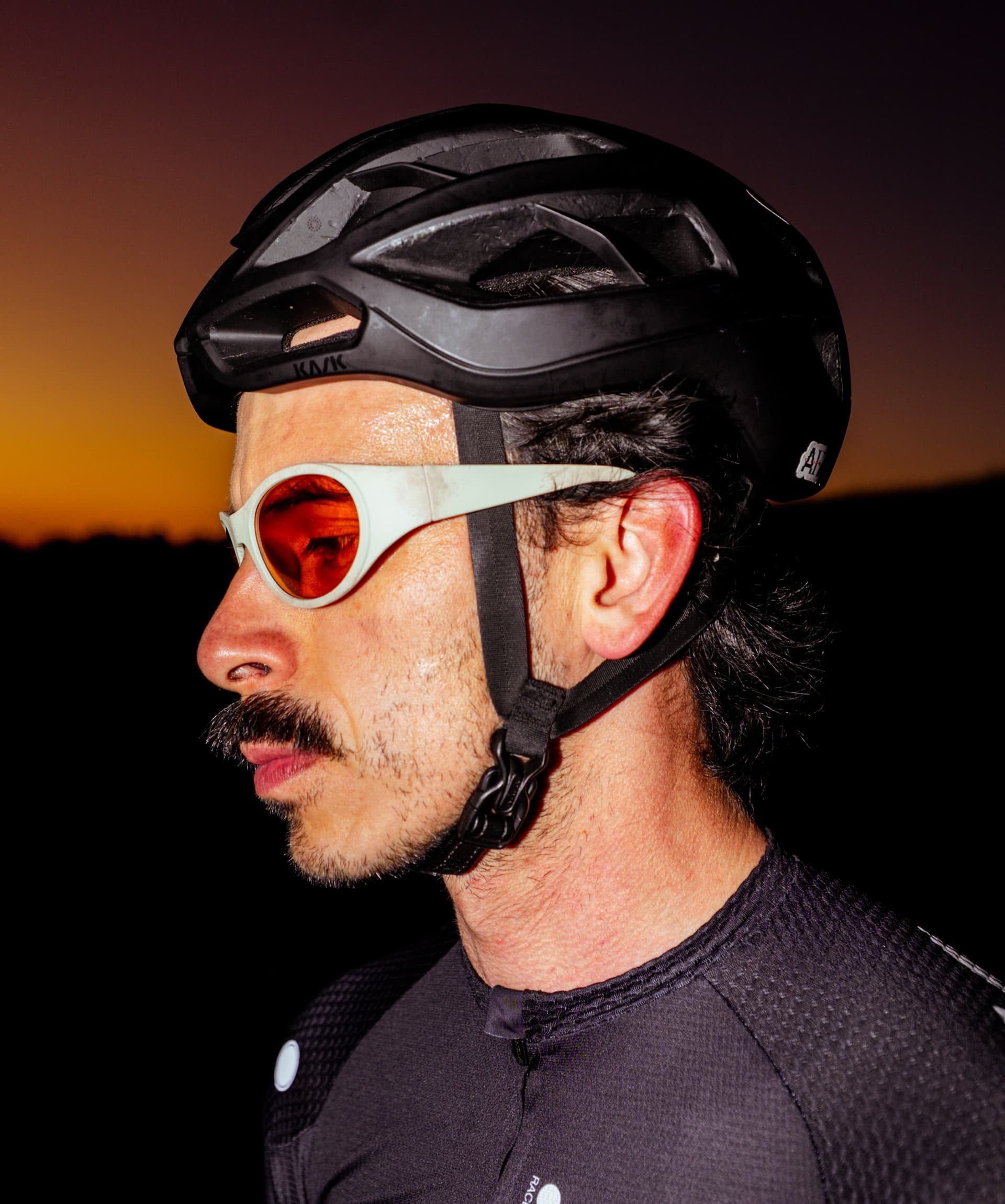 Profile of a man wearing a black cycling helmet and white-framed orange sunglasses, with a sunset in the background.