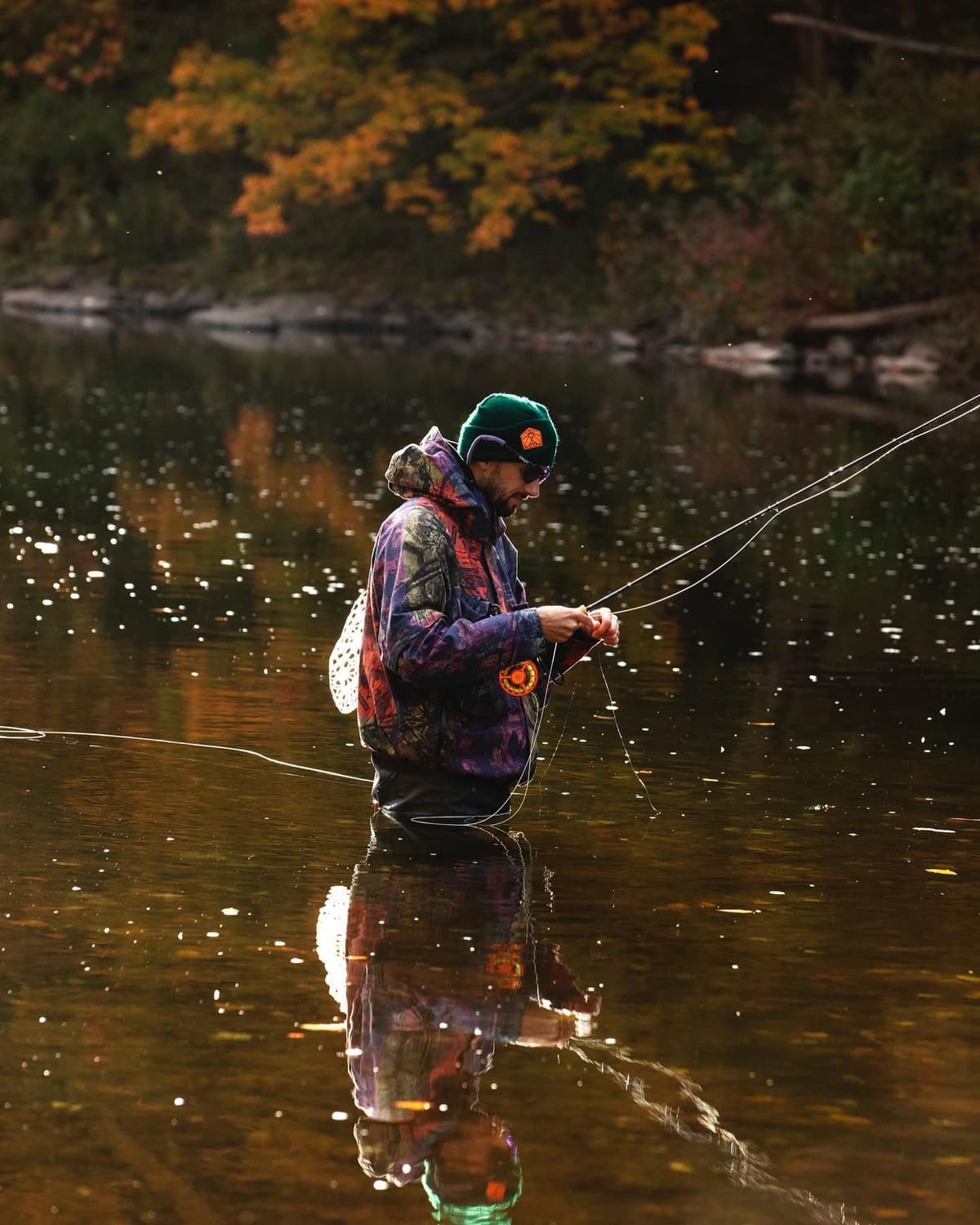 A fly fisherman in a patterned jacket and green beanie stands in a river, his reflection visible, with colorful autumn trees in the background.