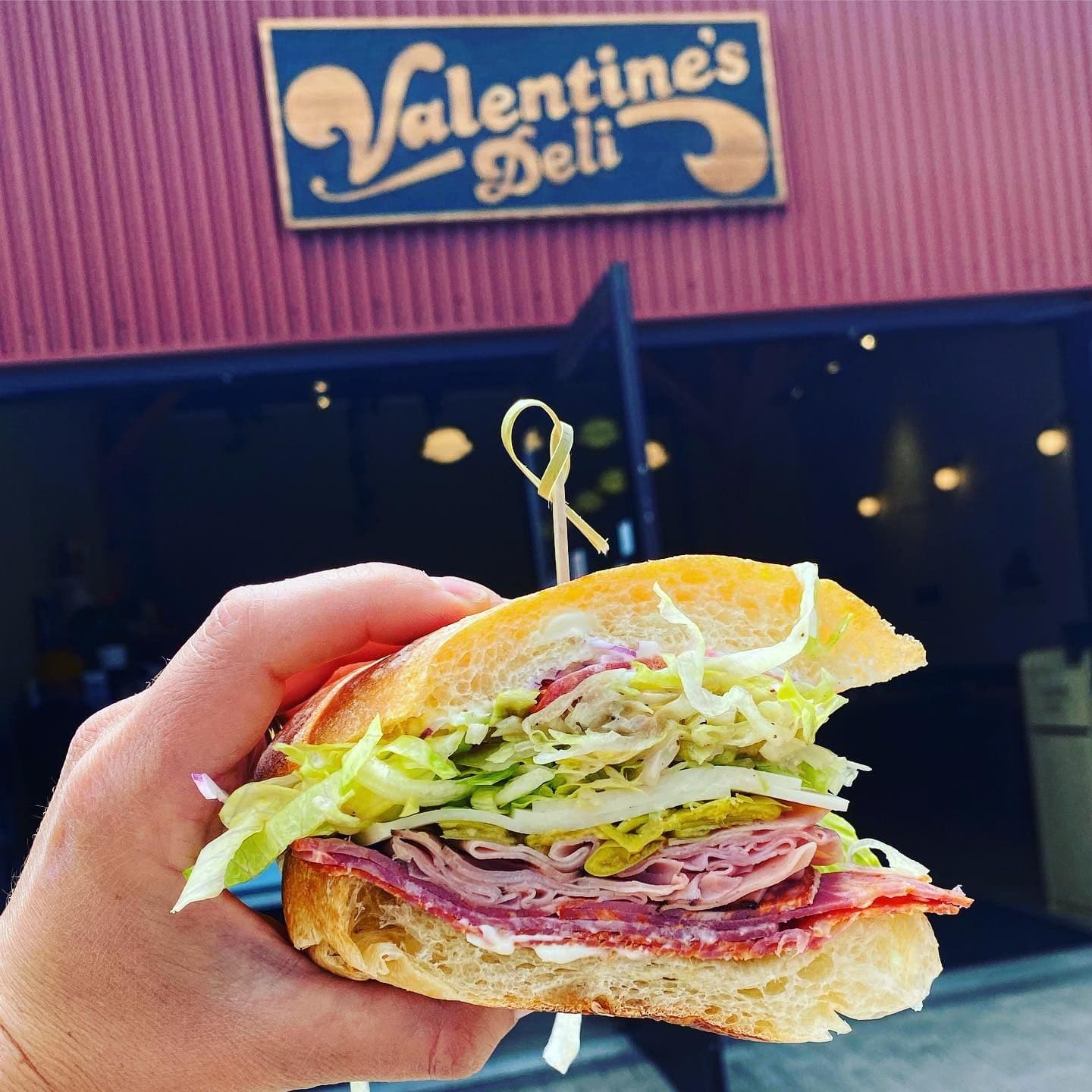 Hand holding a large sub sandwich filled with meat and lettuce, with "Valentine's Deli" sign in the background.