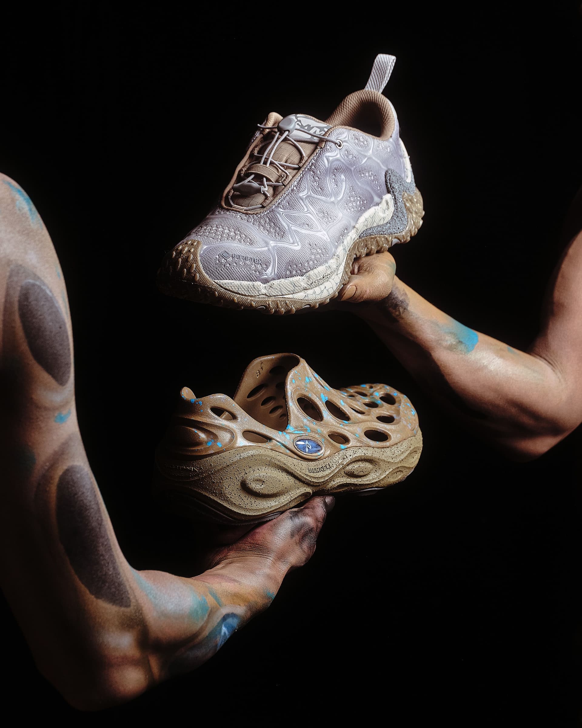 Two arms with abstract earthy and blue paint hold a muddy gray trail shoe and a muddy brown perforated clog.