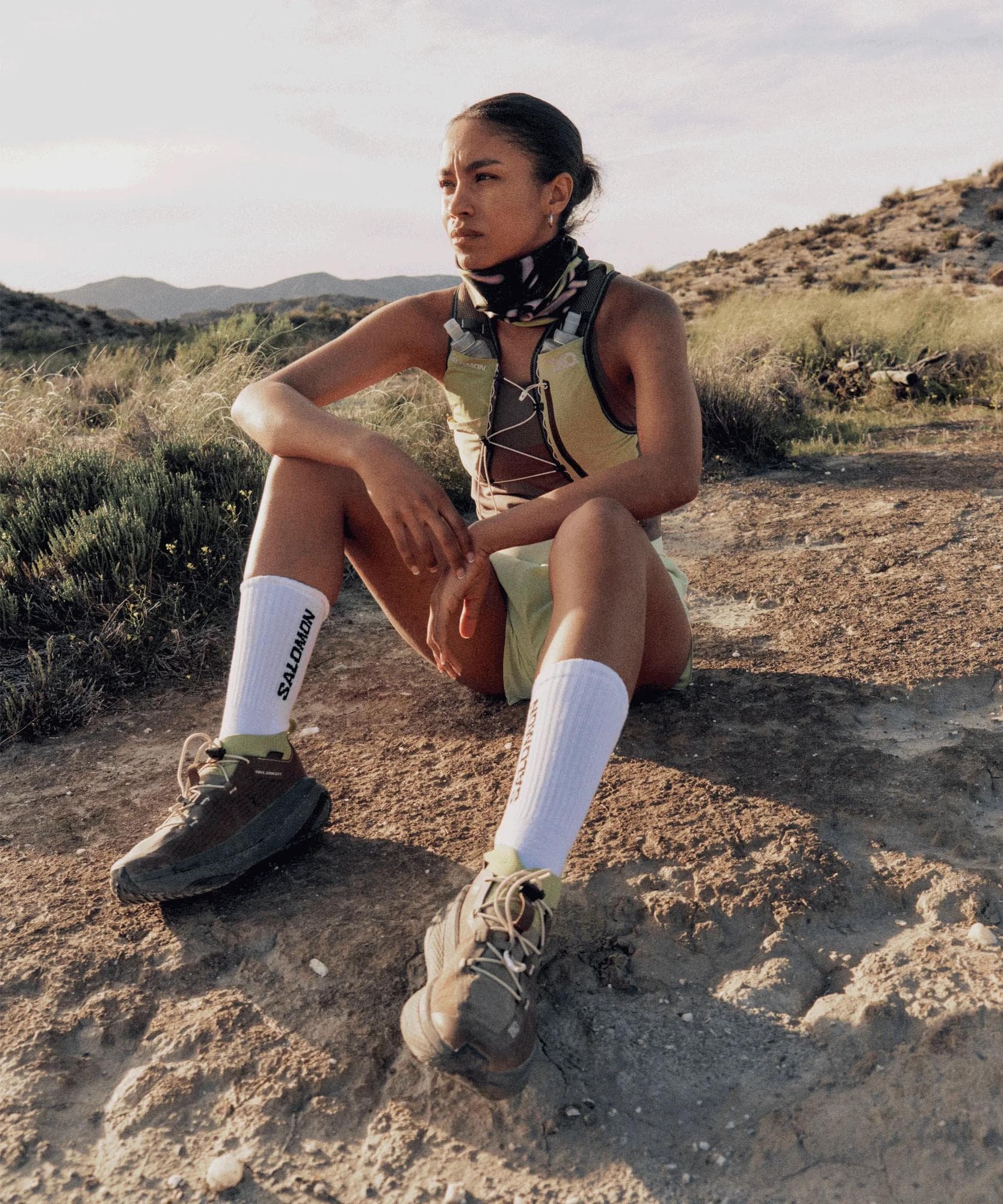 A woman in a hydration vest, patterned neck gaiter, and Salomon trail shoes sits on a dirt path in a desert landscape.