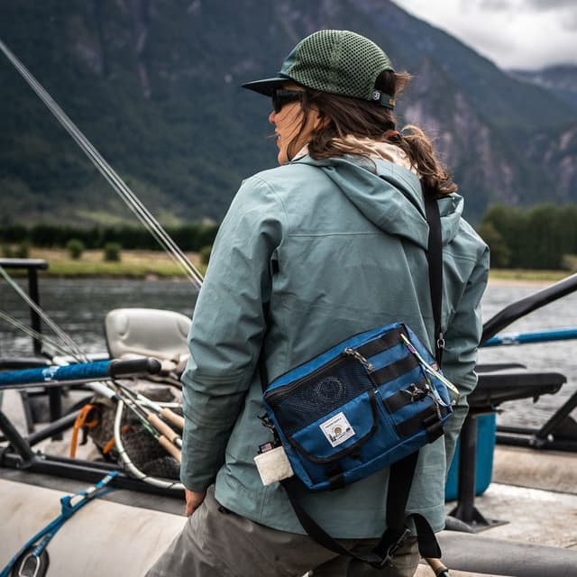 Rear view of a person in a jacket and cap, wearing a blue fishing bag, on a boat with fishing rods and mountains in the background.