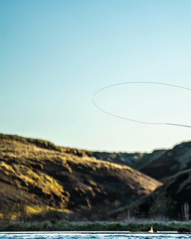 A fly fishing line arcs through the air over a river with hills in the background.