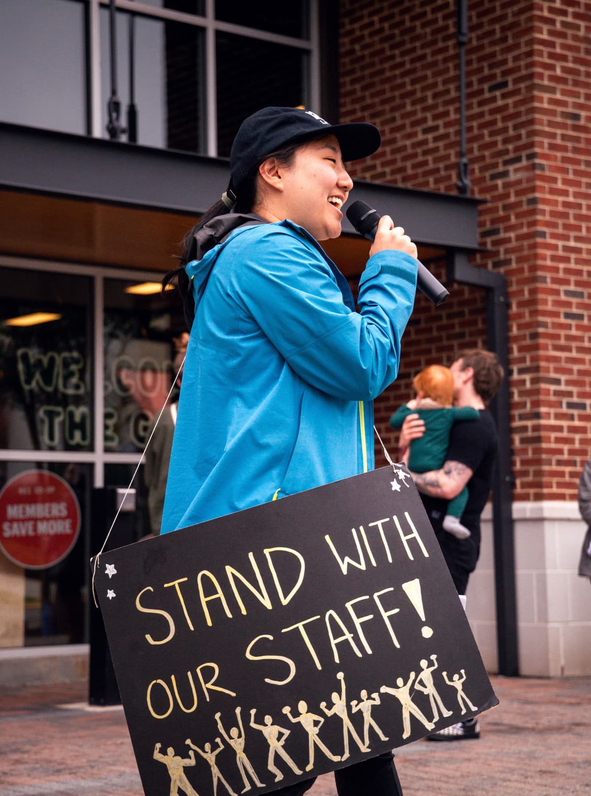 A smiling speaker in a blue jacket holds a microphone and a sign reading "STAND WITH OUR STAFF!" with stick figures.