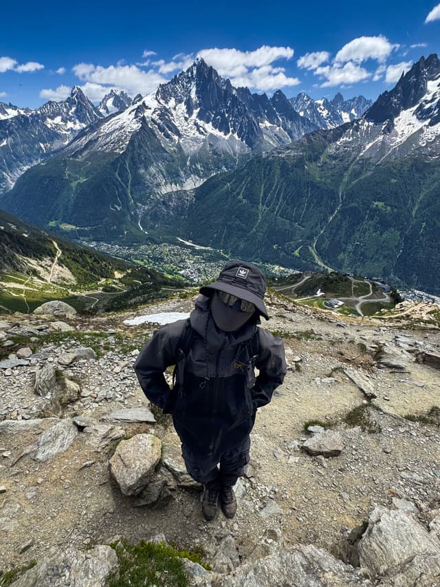 A person in dark hiking gear stands on a rocky mountain path, overlooking snow-capped peaks and a valley town below.