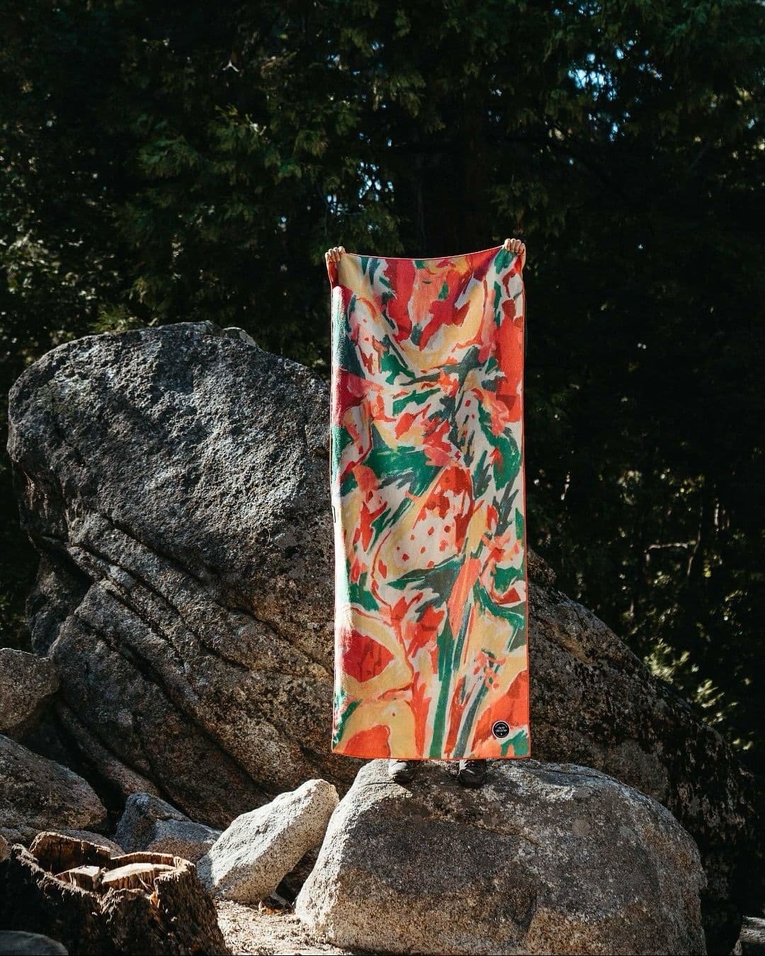 A vibrant red, green, and yellow abstract patterned towel held vertically in a rocky outdoor setting.