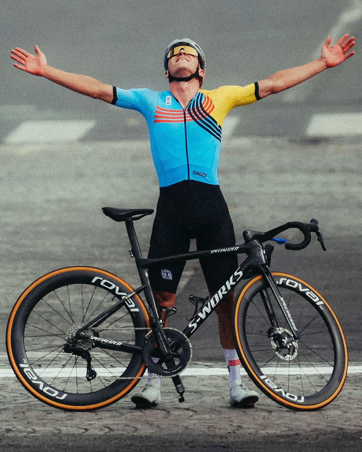 A male cyclist in a blue, red, yellow, and black jersey celebrates with outstretched arms next to a black road bike.