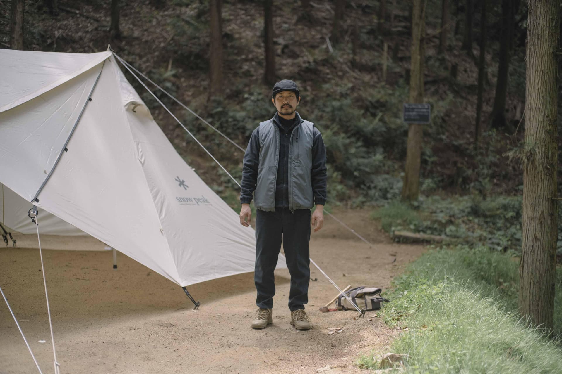 A man in a black cap and vest stands beside a white Snow Peak tent in a forest clearing.