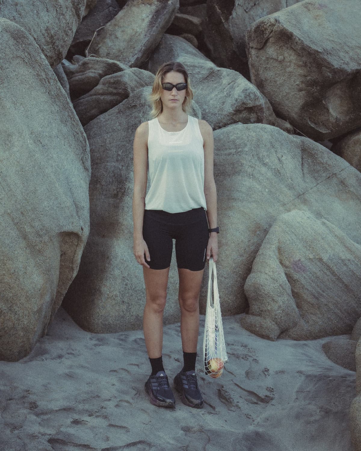 A woman in a white tank top, black shorts, and sunglasses stands among large rocks, holding a string bag with an apple.
