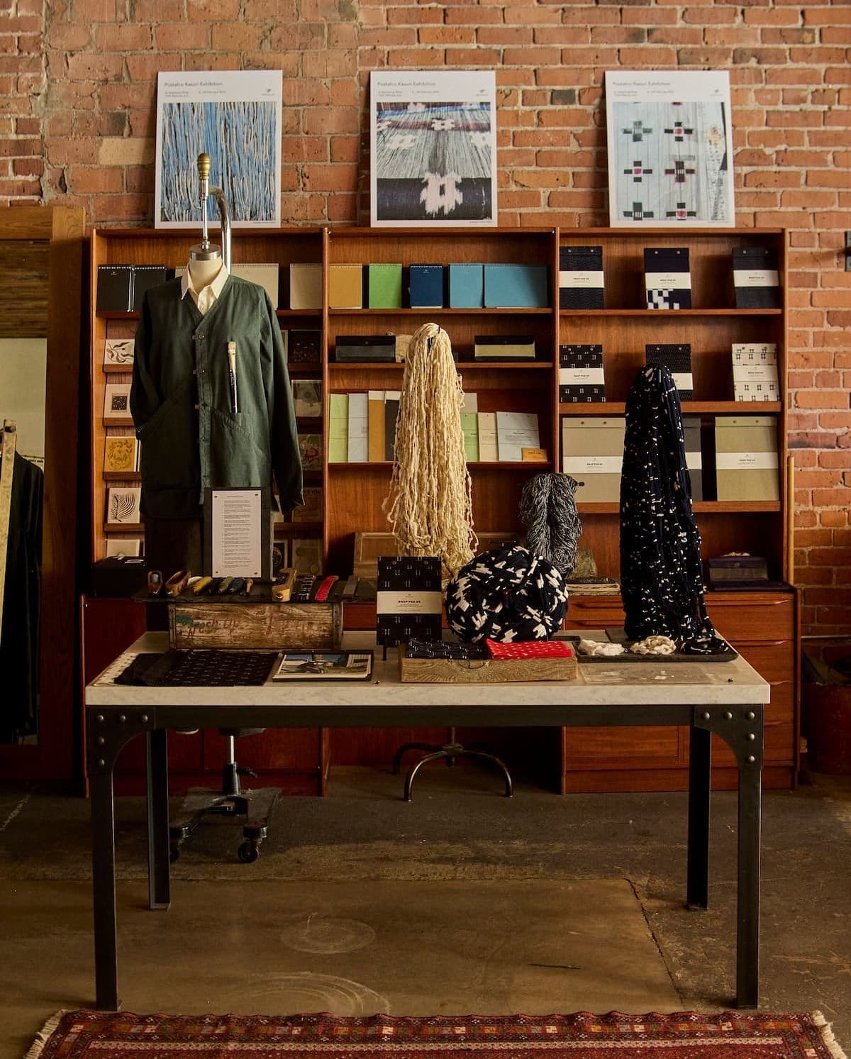 A curated display of textiles, yarns, and artisan goods on a table in a shop with a brick wall and wooden shelves.