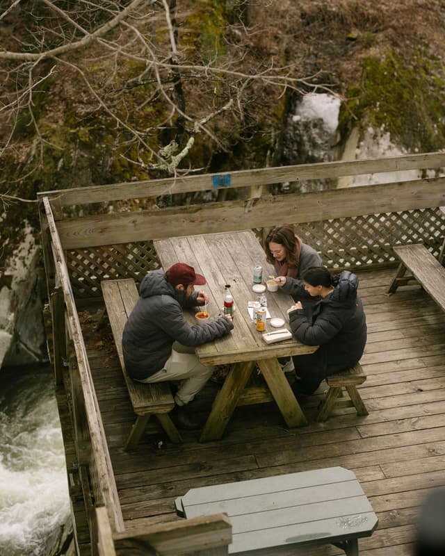 Three people eat at a picnic table on a wooden deck overlooking a rushing river and forested hillside.