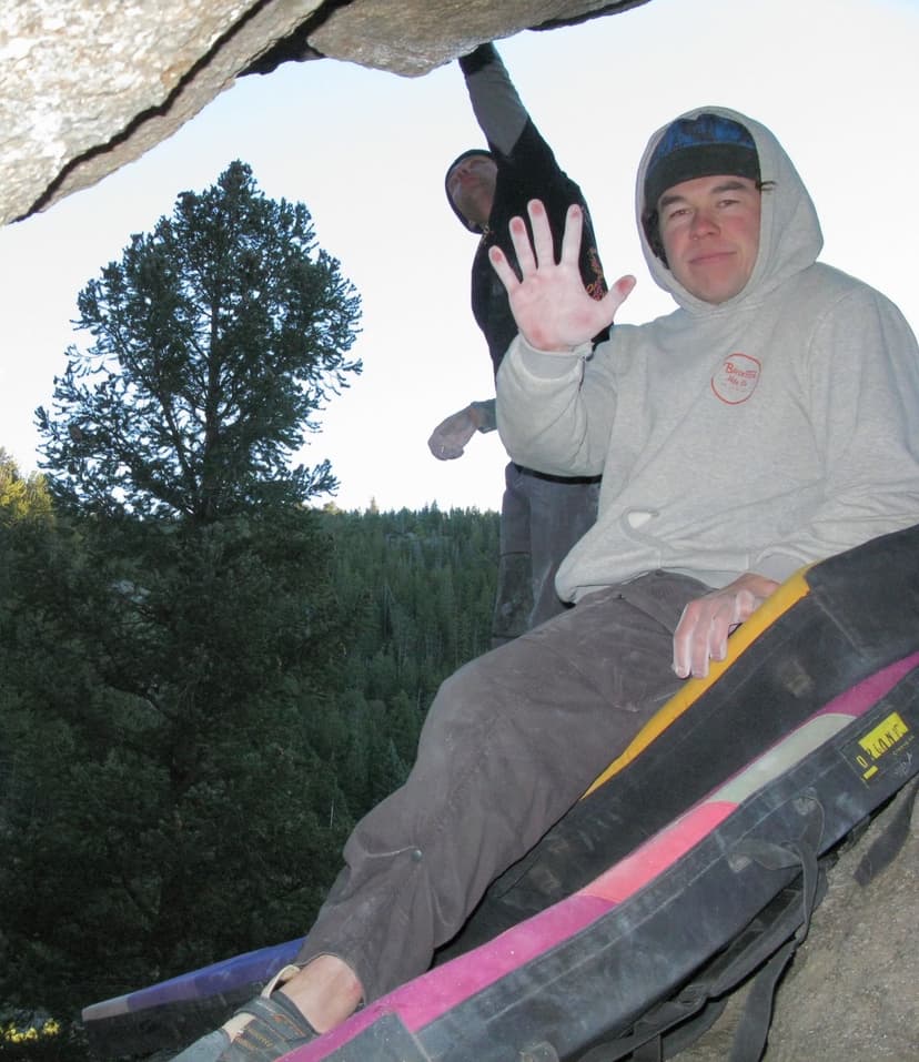 A boulderer sits on crash pads, holding up a chalky hand, with another climber on a rock overhang and a forest in the background.