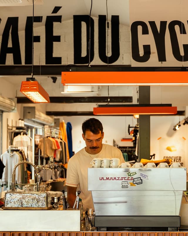 A man with a mustache operates an espresso machine behind a café counter, with clothing and "CAFÉ DU CYCL" visible in the background.