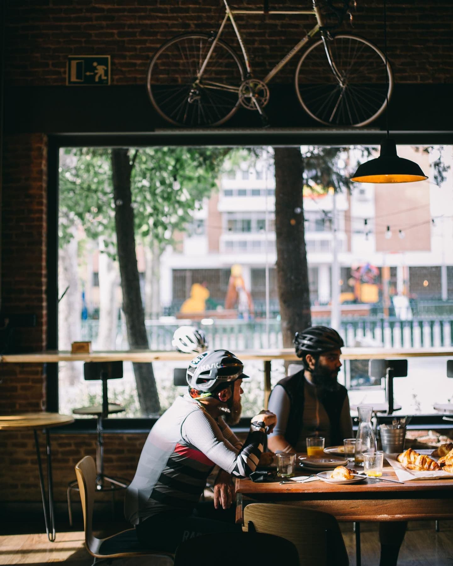 Two cyclists in helmets eat pastries and drinks at a cafe table. A bicycle hangs from the ceiling above them, with a city view outside a large window.