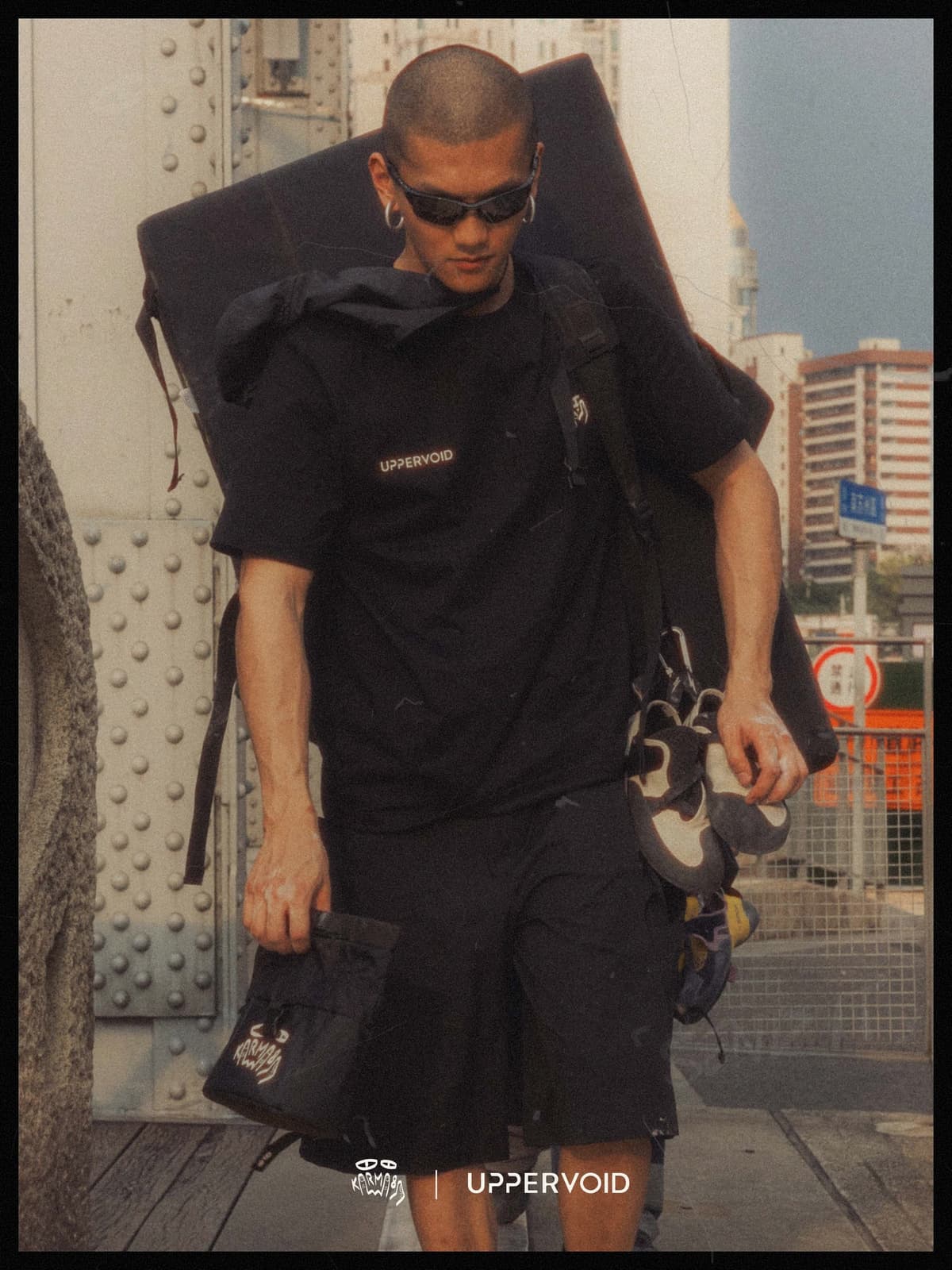 A man with a buzz cut and sunglasses walks in an urban setting, carrying a bouldering crash pad, climbing shoes, and a chalk bag, wearing a black "UPPERVOID" t-shirt and shorts.